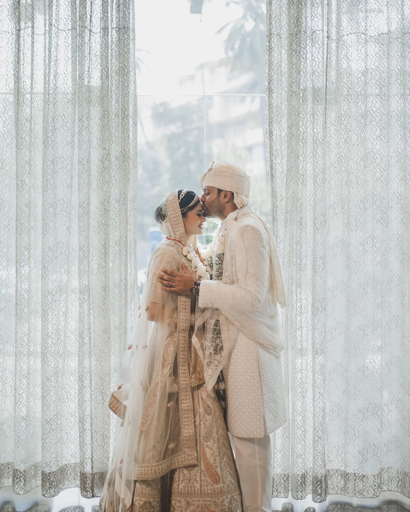 Bride and groom sharing a quiet kiss against a sheer backlit curtain, wedding day