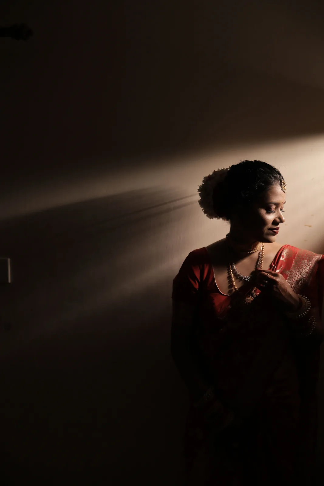 Bride in a red saree lit by a single shaft of window light in a dark room