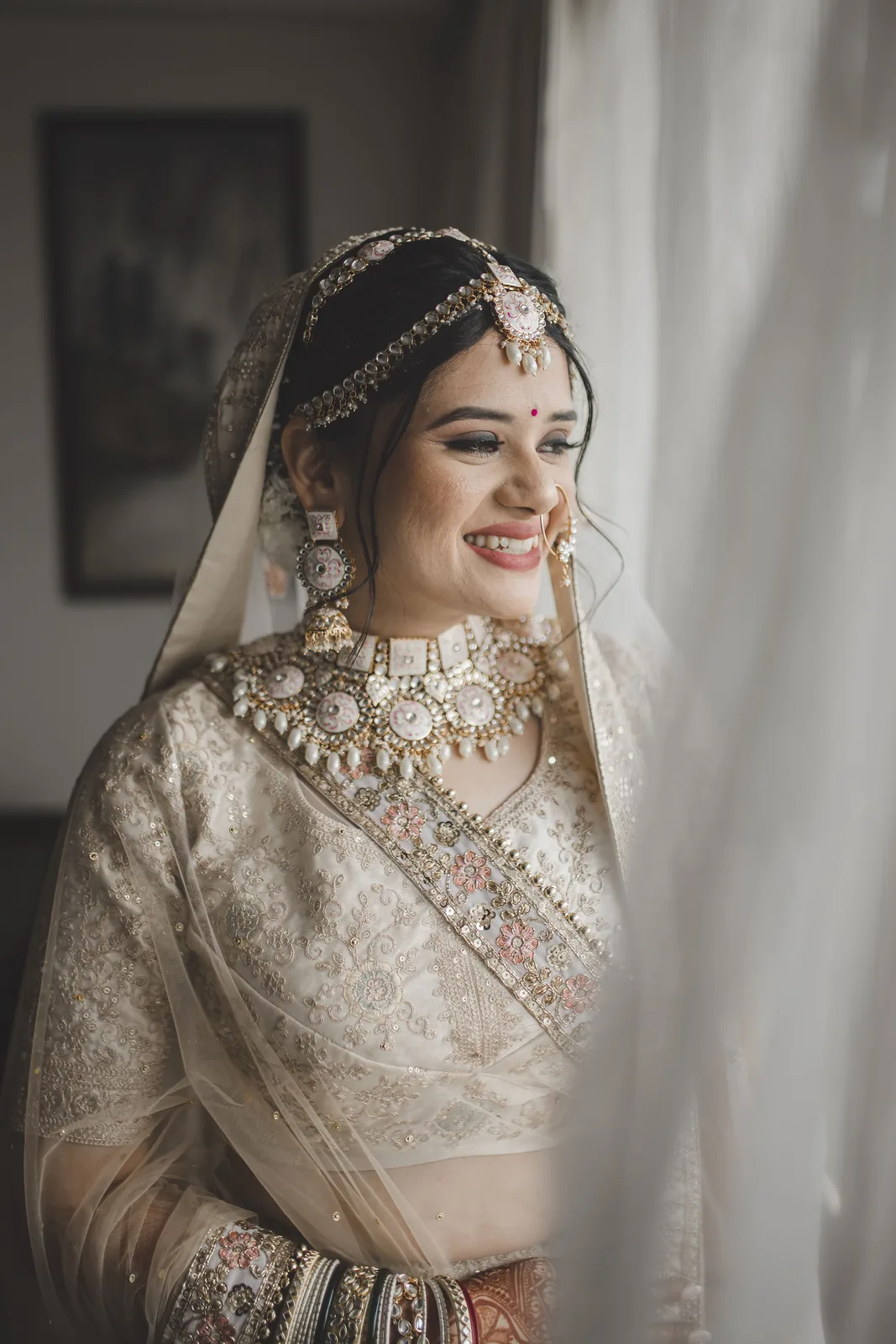Bride in ivory lehenga smiling at a sheer-curtained window