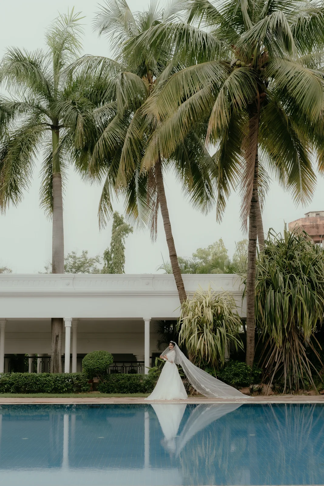 Bride with a long veil at a colonnaded pool between palm trees