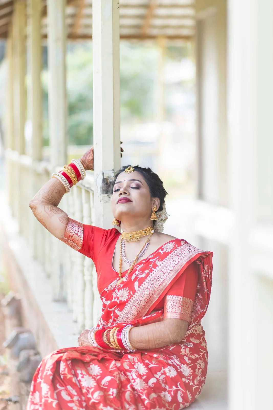 Bride in a red saree resting against a verandah pillar